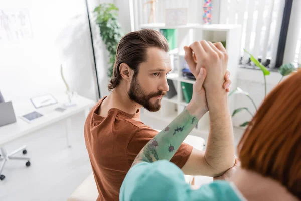 Розмитий фізіотерапевт вивчає руку і лікоть бородатого чоловіка під час призначення в реабілітаційному центрі — стокове фото