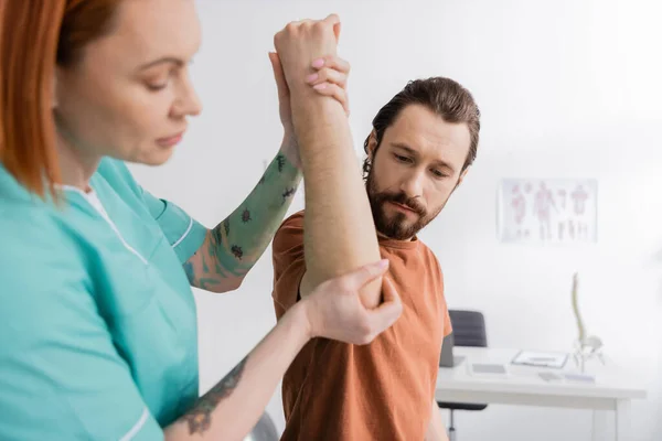 Tätowierter Physiotherapeut beugt Arm eines bärtigen Mannes vor, während er verletzten Ellbogen im Sprechzimmer untersucht — Stockfoto