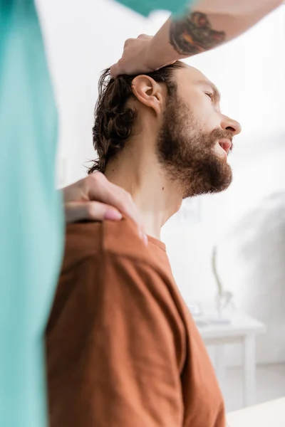 Especialista en rehabilitación borrosa estirando el cuello del paciente barbudo mientras hace diagnósticos en la sala de consulta - foto de stock