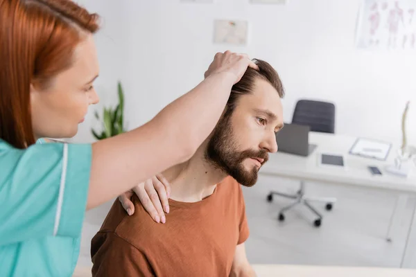 Розмитий фізіотерапевт, який вивчає бородатого чоловіка з травмованою шиєю в консалтинговій кімнаті — стокове фото