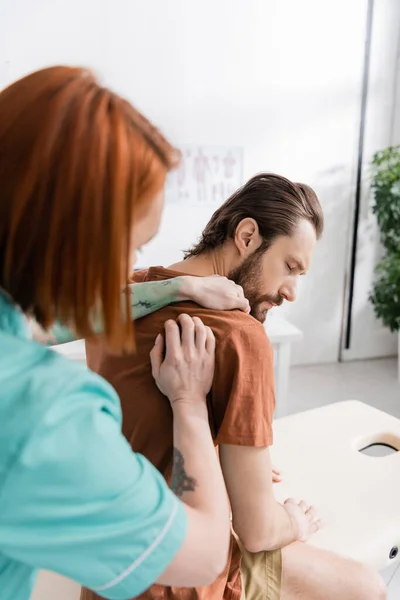 Розмитий хіропрактик вивчає болюче плече пораненого чоловіка в консалтинговій кімнаті — стокове фото