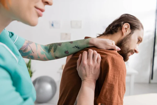Tattooed physiotherapist touching injured shoulder of bearded man in rehabilitation center — Stock Photo