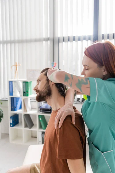 Боковой вид татуированного мануального терапевта, осматривающего болезненную шею бородатого человека в кабинете консультанта — стоковое фото