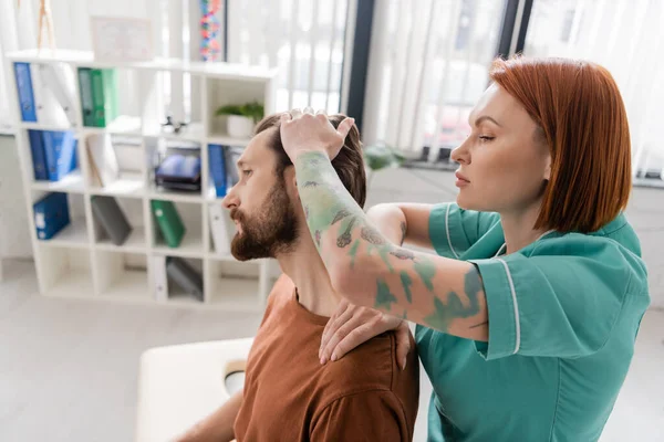 Боковой вид на татуированного физиотерапевта, растягивающего шею бородатого мужчины во время осмотра в реабилитационном центре — стоковое фото