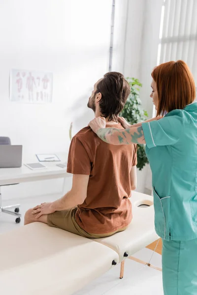 Рудий остеопат торкається болючої спини людини, що сидить на масажному столі в консалтинговій кімнаті — стокове фото