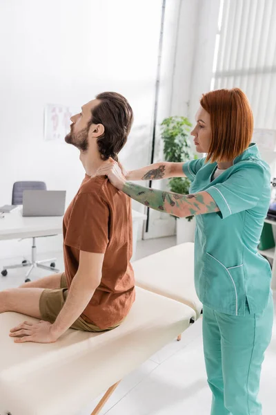 Vue latérale d'un ostéopathe examinant le dos d'un homme barbu assis sur une table de massage dans une salle de consultation — Photo de stock