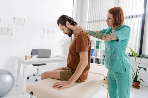 Vue latérale du kinésithérapeute rousse examinant le dos blessé d'un homme barbu assis sur une table de massage dans une salle de consultation — Photo de stock