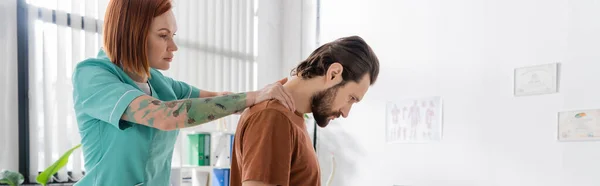 Vista lateral del quiropráctico pelirrojo examinando la espalda lesionada del paciente barbudo en el centro de rehabilitación, pancarta - foto de stock