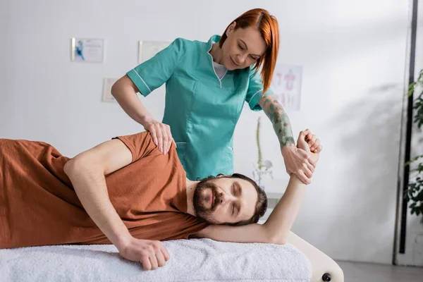 Redhead rehabilitologist massaging painful shoulder of bearded man with closed eyes in consulting room — Stock Photo