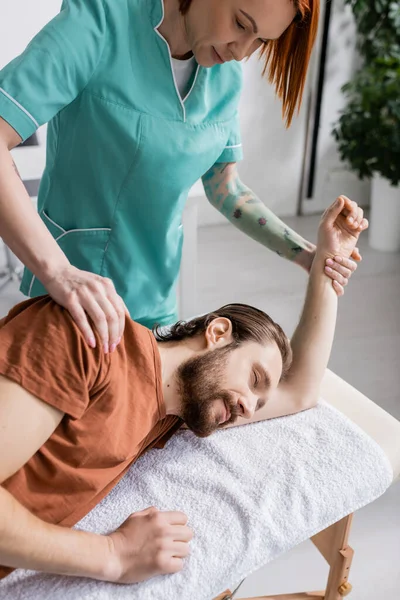 Fisioterapeuta tatuado masajeando hombro lesionado de hombre barbudo en centro de rehabilitación - foto de stock