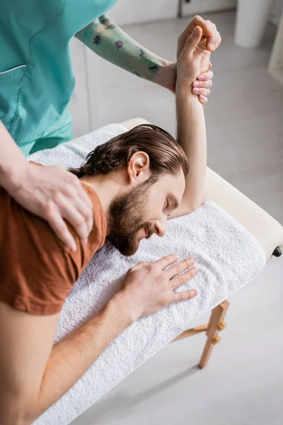 Tattooed chiropractor massaging shoulder of injured man during manual therapy in consulting room — Stock Photo
