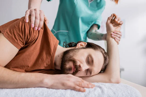 Физиотерапевт массирует травмированное плечо бородатого мужчины с закрытыми глазами в реабилитационном центре — стоковое фото