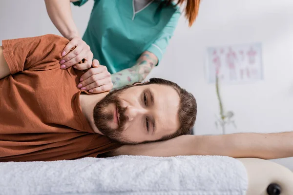 Татуированный физиотерапевт массирует больное плечо бородатого мужчины в реабилитационном центре — стоковое фото