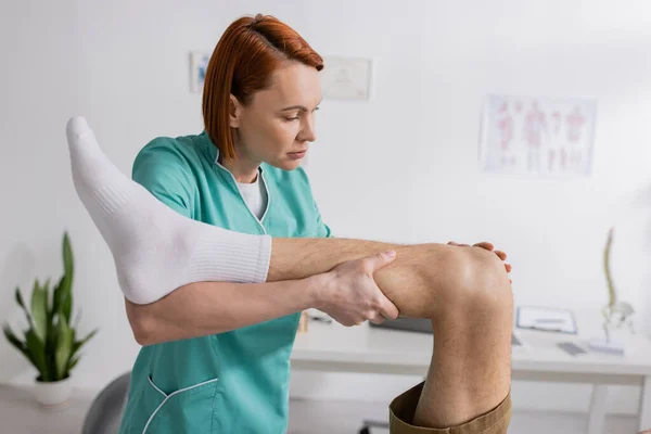 Терапевт-рыжеволосый, работающий с травмой ноги человека в реабилитационном центре — стоковое фото