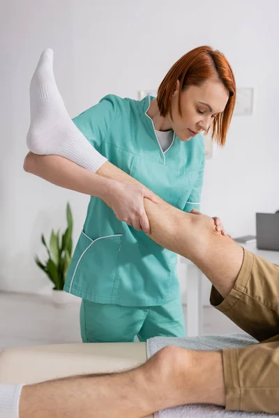 Fisioterapeuta estirar la pierna del hombre lesionado durante la terapia de rehabilitación en la sala de consulta - foto de stock