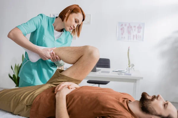 Fisioterapeuta trabalhando com perna ferida do homem deitado na mesa de massagem em consultório — Fotografia de Stock