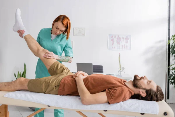 Terapeuta manual ruiva massageando perna dolorosa do homem barbudo no centro de reabilitação — Fotografia de Stock