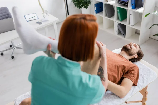 Verschwommene manuelle Therapeutin beugt verletztes Bein des Mannes vor, während sie Schmerzlindermassage im Reha-Zentrum macht — Stockfoto