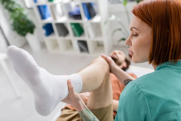 Pelirroja fisioterapeuta flexión pierna de hombre durante la terapia de recuperación en la sala de consulta - foto de stock