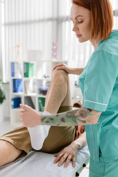 Quiropráctico tatuado haciendo masaje de piernas al hombre lesionado durante el tratamiento en la sala de consulta - foto de stock