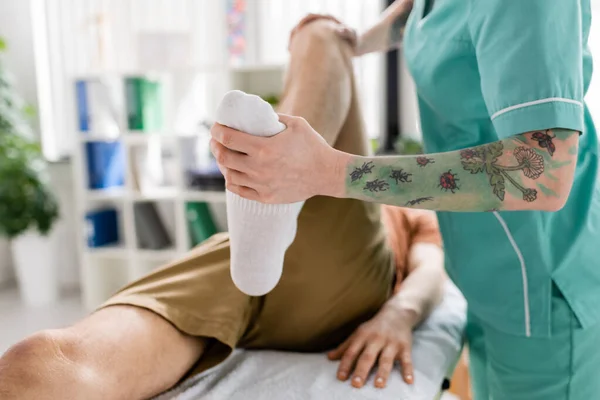 Visão recortada do quiroprático tatuado que flexiona a perna do paciente durante o tratamento no centro de reabilitação — Fotografia de Stock