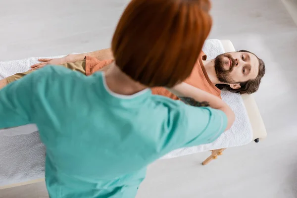 Vue grand angle de kinésithérapeute rousse faisant massage soulagement de la douleur à l'homme blessé dans la salle de consultation — Photo de stock