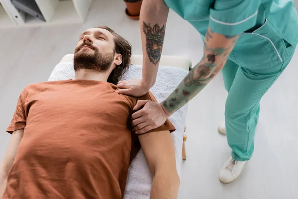 Quiropráctico tatuado haciendo masaje en el hombro a hombre barbudo herido acostado en la sala de consulta - foto de stock