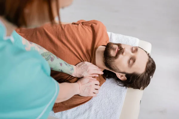 Высокий угол обзора бородатого мужчины с закрытыми глазами рядом с размытым физиотерапевтом, делающим массаж плеч в консультационной комнате — стоковое фото