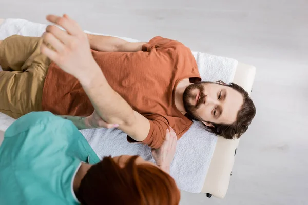 Вид зверху розмитого мануального терапевта масажу пораненої руки бородатого чоловіка в реабілітаційному центрі — стокове фото