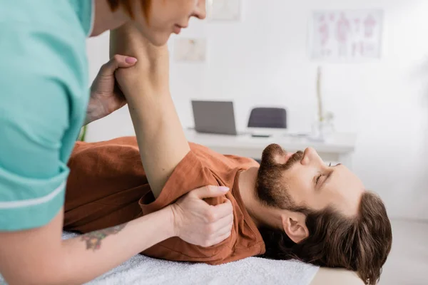 Vue latérale du spécialiste de la réadaptation massant le bras douloureux de l'homme barbu dans la salle de consultation — Photo de stock