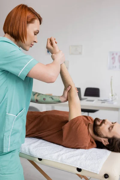 Redhead physiotherapist stretching painful arm of bearded man lying on massage table in rehabilitation center — Stock Photo