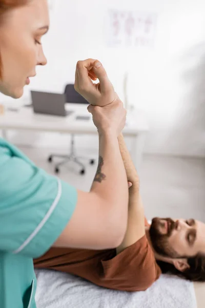 Fisioterapeuta estiramiento brazo lesionado del hombre borroso durante la rehabilitación en el hospital - foto de stock