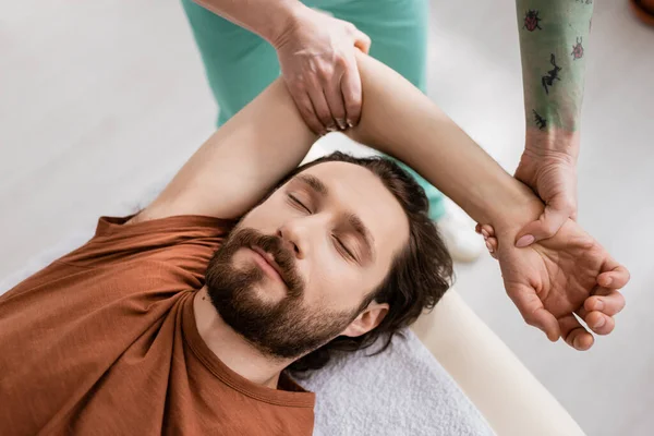 Vista superior del hombre barbudo con los ojos cerrados cerca de fisioterapeuta haciendo masaje de brazo durante la rehabilitación en la clínica - foto de stock