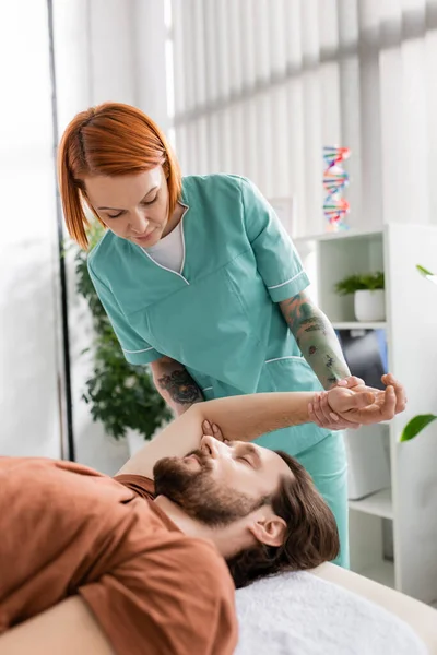 Terapeuta manual ruiva flexionando braço doloroso do homem barbudo durante o tratamento de reabilitação no hospital — Fotografia de Stock