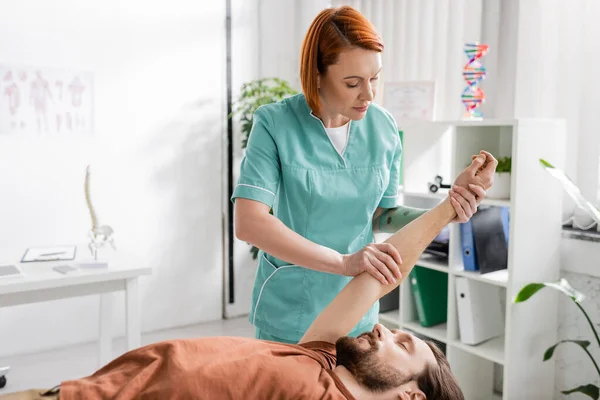 Рудий хіропрактик розтягує руку пораненого чоловіка під час проведення масажу полегшення болю в консалтинговій кімнаті — стокове фото