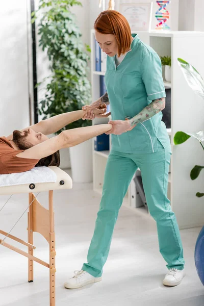 Fisioterapeuta tatuado estirando los brazos del hombre barbudo durante el tratamiento de rehabilitación en la clínica - foto de stock