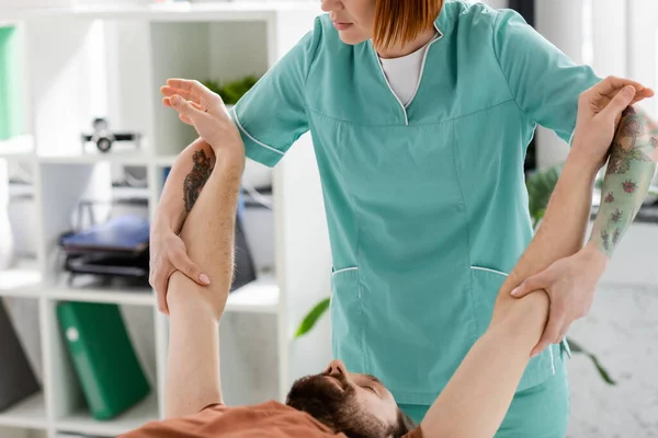Manuelle Therapeutin streckt Arme eines bärtigen Mannes während der Rehabilitation in der Klinik — Stockfoto
