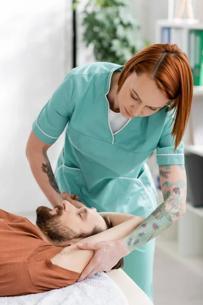 Redhead chiropractor massaging arm of bearded man during recovery therapy in consulting room — Stock Photo