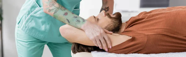 Татуированный физиотерапевт, делающий обезболивающий массаж раненому человеку в реабилитационном центре, баннер — стоковое фото