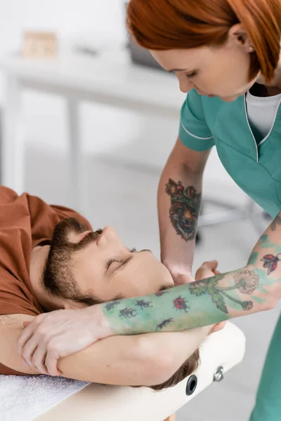Татуированный остеопат массирует травмированную руку бородатого мужчины в центре реабилитации — стоковое фото