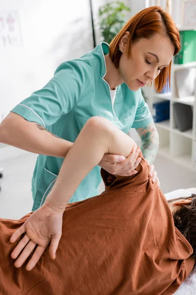 Fisioterapeuta pelirroja haciendo masaje de brazo y hombro a paciente en centro de rehabilitación - foto de stock
