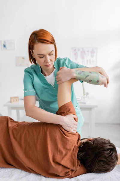 Brazo de flexión del fisioterapeuta tatuado del paciente durante la terapia de alivio del dolor en el centro de rehabilitación - foto de stock