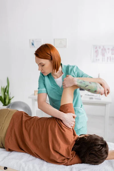 Рыжеволосый физиотерапевт, делающий обезболивающий массаж на поврежденной руке человека в кабинете консультанта — стоковое фото