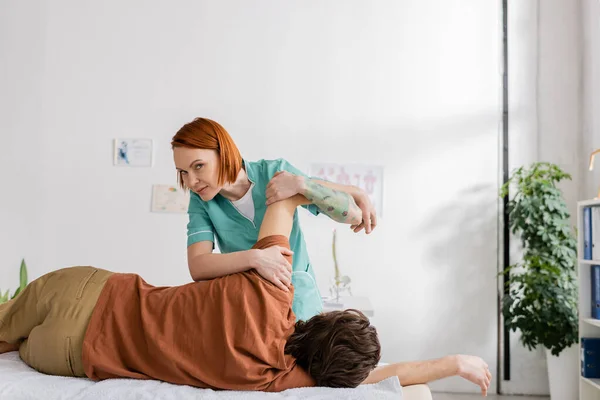 Chiropratico rossa guardando la fotocamera mentre flette il braccio ferito dell'uomo durante il massaggio antidolorifico nella sala di consultazione — Foto stock