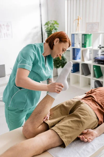 Rotschopf-Osteopath beugt Bein eines Mannes, der im Reha-Zentrum auf Massagetisch liegt — Stockfoto