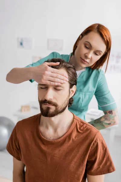 Bearded man with closed eyes near redhead physiotherapist doing diagnostics in rehabilitation center — Stock Photo