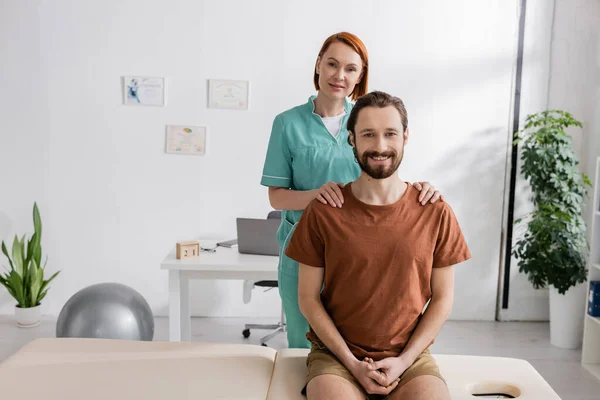 Fisioterapista felice toccare le spalle di uomo barbuto seduto sul tavolo da massaggio in sala di consultazione — Foto stock