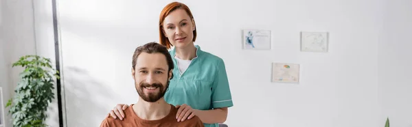 Радісний фізіотерапевт і бородатий чоловік посміхається на камеру в реабілітаційному центрі, банер — стокове фото