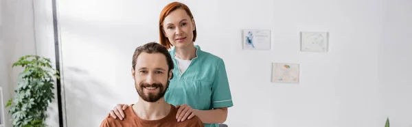 Fisioterapeuta alegre y hombre barbudo sonriendo a la cámara en el centro de rehabilitación, pancarta - foto de stock