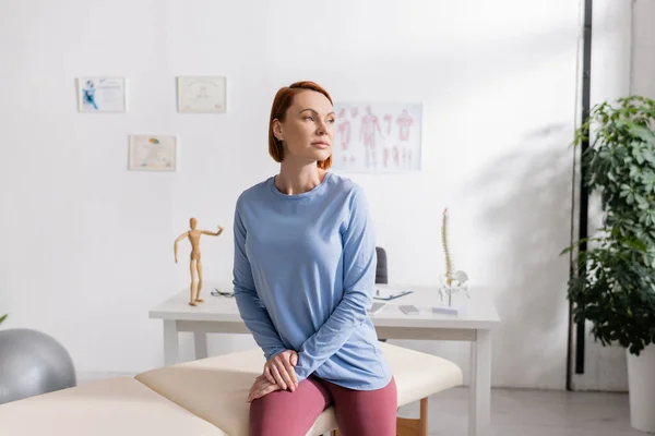 Redhead woman sitting on massage table in rehab center and looking away — Stock Photo